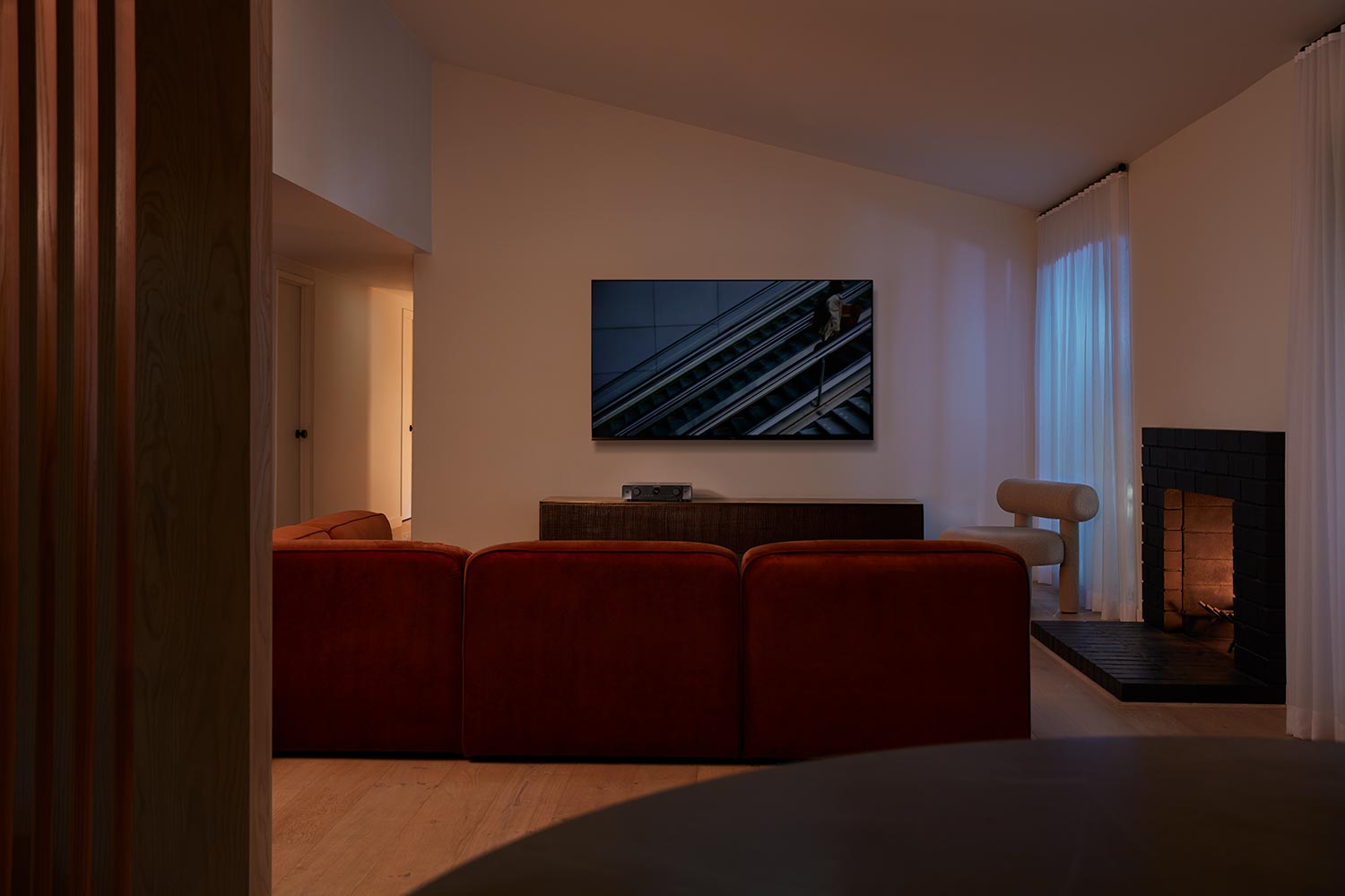 A cozy living room with a red sofa facing a mounted TV, a lit fireplace on the right, and soft white curtains.